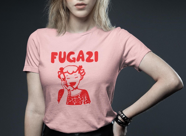 May include: A pink t-shirt with a red graphic of a girl on the phone and the word "FUGAZI" above her.