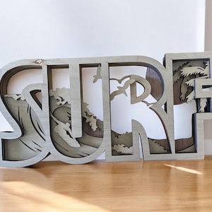 May include: A wooden wall decoration with the word "SURF" in layered, cutout letters. The "S" features a wave with a surfer, the "U" has a palm tree, and the "R" has a cloud.