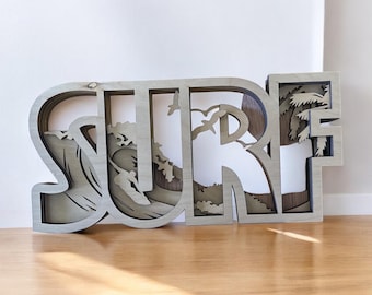 May include: A wooden wall decor with the word "SURF" in layered, cutout letters. The "S" features a wave with a surfer, the "U" has a palm tree, and the "R" has a cloud.