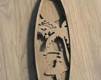 May include: A wooden surfboard-shaped wall art piece. The design features a surfer, palm trees, and the ocean, all cut out from the wood. The art is light brown with a dark brown border, creating a layered effect.