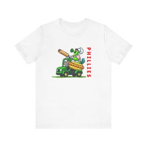 Könnte beinhalten: Weißes T-Shirt mit einer grünen Comicfigur, die ein grünes Fahrzeug mit einem Hotdog im Heck fährt. Die Figur trägt eine Kochmütze und eine Sonnenbrille. Der Text "PHILLIES" ist in Rot auf der rechten Seite des Shirts gedruckt.