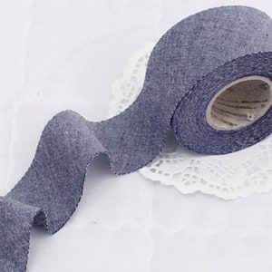 May include: A roll of blue denim ribbon with a scalloped edge. The ribbon is on a white doily.
