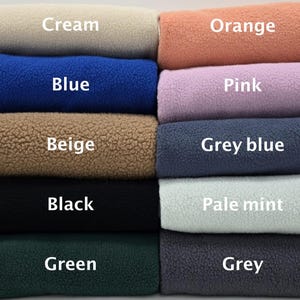 May include: A selection of fleece fabric rolls in various colours: cream, orange, blue, pink, beige, grey blue, black, pale mint, green, and grey. Each roll is labelled with its colour name in white text.