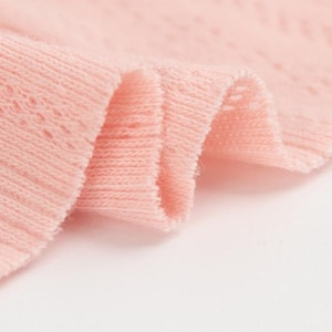 May include: Close-up of a section of soft, light pink knit fabric. The fabric is folded, revealing layers and textures. The knit has a delicate openwork pattern, adding visual interest to the textile. The overall appearance is smooth and gentle.