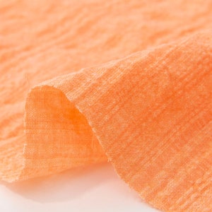 May include: Close-up of a soft, peach-colored fabric with a textured weave. The fabric is folded and draped, showing its texture and color.
