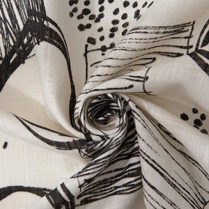 Puede incluir: Primer plano de una tela color crema con un diseño abstracto negro. El diseño presenta pinceladas, puntos y elementos lineales, creando un patrón dinámico. La tela está doblada, mostrando textura y profundidad.