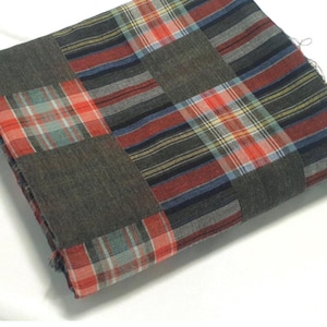 May include: A folded patchwork textile. It combines solid grey squares with plaid squares. The plaid design includes red, blue, and green stripes on a white background. The textile's construction is visible due to its folded state.