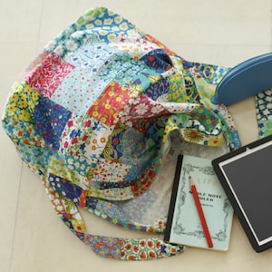 May include: A patchwork tote bag with a floral print in shades of blue, green, red, and yellow. The bag has a long strap and is filled with a notebook, pencil, and tablet.