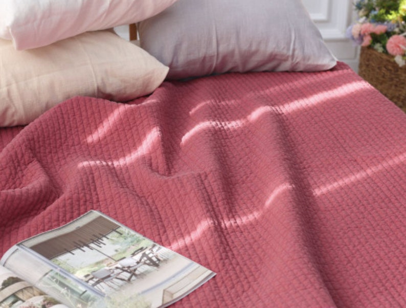 Puede incluir: Una suave manta de tiro acolchada de color rosa con una revista encima. La manta est&aacute; hecha de un tejido texturizado y tiene un brillo sutil.