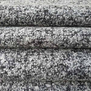 May include: Close-up of a stack of gray and black tweed fabric. The fabric has a textured appearance with a woven pattern. The colors are a mix of light gray, dark gray, and black, creating a speckled effect.