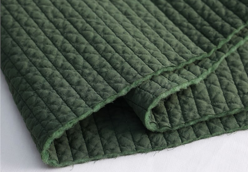 Puede incluir: Primer plano de una tela acolchada de color verde oscuro con un patr&oacute;n de rombos. La tela est&aacute; doblada sobre s&iacute; misma, mostrando la textura suave.