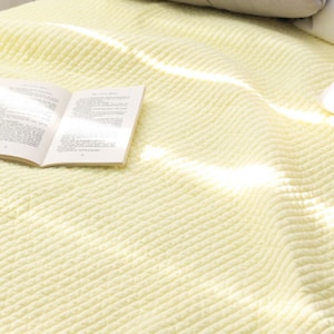 Puede incluir: Una manta suave de color amarillo p&aacute;lido con textura acanalada. La manta est&aacute; doblada y tiene un libro encima.
