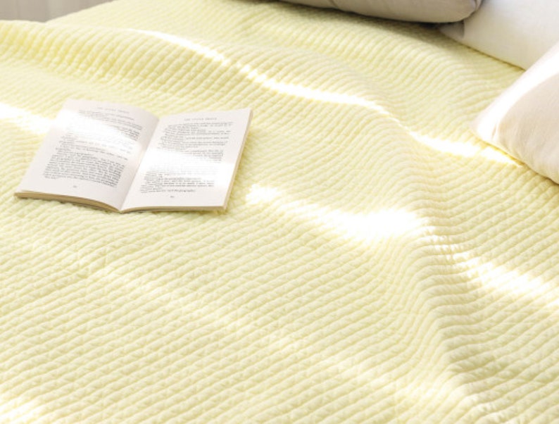 Puede incluir: Una manta suave de color amarillo p&aacute;lido con textura acanalada. La manta est&aacute; doblada y tiene un libro encima.