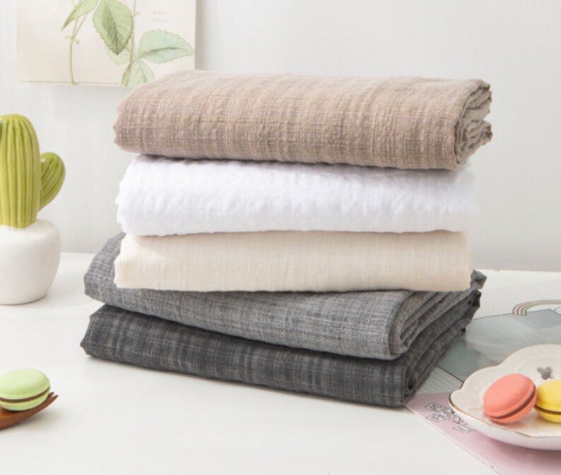 May include: A stack of four fabric swatches in different colors and textures. The swatches are folded and arranged in a pyramid shape. The top swatch is a light brown color, the second swatch is white, the third swatch is a light beige color, and the bottom swatch is a dark gray color.