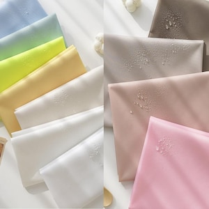 May include: A collection of fabric swatches in various colours, including light blue, yellow, beige, white, and pink. The fabric is smooth and has a subtle sheen. Water droplets are visible on the surface of the fabric, indicating that it is water-resistant.