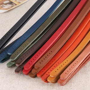 Puede incluir: Una colección de correas de cuero coloridas en varios tonos, incluyendo negro, azul, verde, rojo y naranja. Cada correa tiene una serie de agujeros espaciados uniformemente, lo que sugiere que están diseñadas para la fijación o el ajuste. Las correas están dispuestas en diagonal.