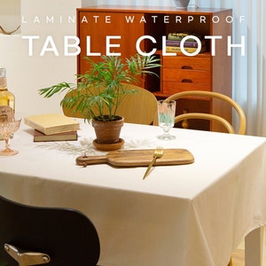 May include: White laminated waterproof tablecloth on a table with a potted plant, books, a wine bottle, a glass, and a fork. The text "Laminate Waterproof Table Cloth" is displayed above the table.