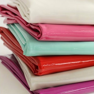 Puede incluir: Una pila de cinco telas de vinilo de diferentes colores. Las telas están dobladas y apiladas una encima de la otra. Los colores son blanco, rosa, azul claro, rojo y morado.