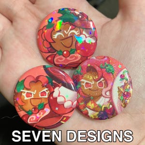 May include: Three round, colorful pinback buttons featuring cartoon characters with strawberry and cherry themes. The buttons have a holographic effect and the text "SEVEN DESIGNS" is at the bottom.