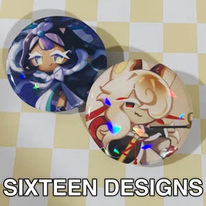 May include: Two round pinback buttons with cartoon character designs. One button features a character in blue and white, the other in tan and red. The text "SIXTEEN DESIGNS" is printed in white at the bottom.