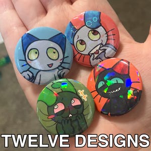 May include: Four round buttons with cartoon cat designs in blue, orange, green, and red. Each button features a unique cat illustration with holographic accents. The text "TWELVE DESIGNS" is at the bottom.