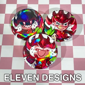 May include: Three round, holographic buttons featuring cartoon character designs in red, white, and green. The buttons have a glossy finish and are arranged on a pink and white checkered background. The text "ELEVEN DESIGNS" is at the bottom.
