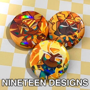 May include: Three round buttons with cartoon character designs in shades of yellow, orange, and brown. The buttons feature a holographic effect and the text "NINETEEN DESIGNS" at the bottom.