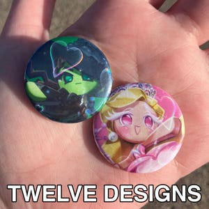 Könnte beinhalten: Zwei runde Buttons mit Cartoon-Charakter-Designs. Ein Button zeigt eine grüne Hexe mit einer herzförmigen Blase, der andere eine blonde Prinzessin mit rosa Akzenten. Der Text "TWELVE DESIGNS" befindet sich unten.