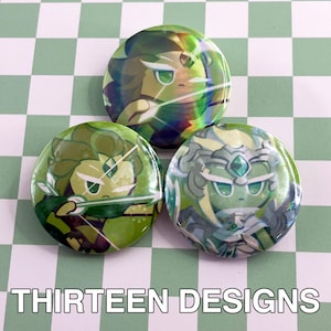 May include: Three round, green and iridescent buttons featuring cartoon characters. The buttons have a glossy finish and are arranged on a green and white checkered surface. The text "THIRTEEN DESIGNS" is at the bottom.