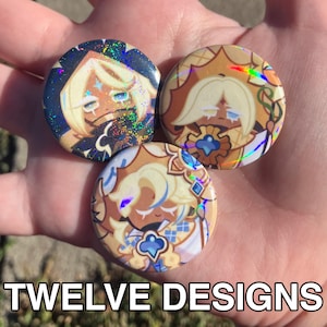 May include: Three round buttons with character illustrations and holographic accents. The buttons feature a light-haired character with various outfits and expressions. The text "TWELVE DESIGNS" is at the bottom.