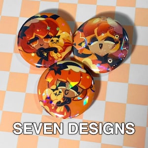 May include: Three round orange buttons with cartoon character designs. The buttons have a holographic effect and feature characters with orange hair and black outfits. The text "SEVEN DESIGNS" is at the bottom.
