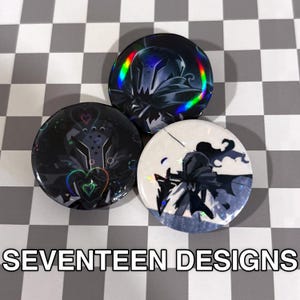 May include: Three round buttons with holographic designs. One button has a black background with a design of hearts and a helmet. Another button has a black background with a design of a helmet. The third button has a white background with a design of a person. The text "SEVENTEEN DESIGNS" is at the bottom.