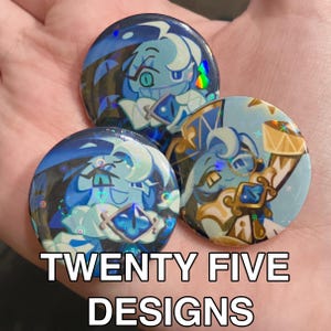 May include: Three round, blue and white character button pins with holographic accents. The pins feature a cartoon character design with the text "TWENTY FIVE DESIGNS" in white block letters.