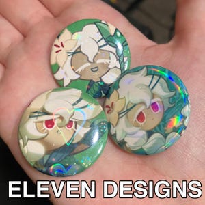 May include: Three round buttons with a holographic effect. Each button features a cartoon character with white hair, red eyes, and a floral design. The text "ELEVEN DESIGNS" is at the bottom.