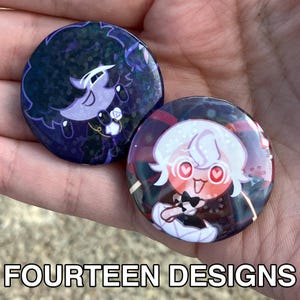 May include: Two round pinback buttons with cartoon character designs. One button has a dark blue background with a purple character. The other button has a red and black background with a white character. The text "FOURTEEN DESIGNS" is at the bottom.
