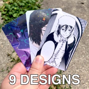 May include: Three rectangular trading cards featuring anime-style illustrations. One card displays a character in a fur-lined hat with a speech bubble, while the others show holographic effects with star and heart patterns. The text 9 DESIGNS is visible.