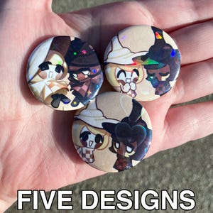 May include: Three circular pin badges with cartoon character designs. The badges feature light tan, brown, and black colours with holographic accents. The text "FIVE DESIGNS" is at the bottom of the image.