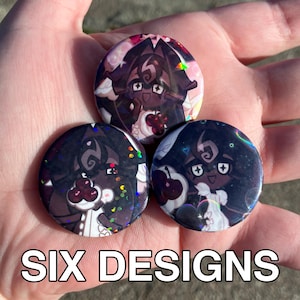 May include: Three round pinback buttons with cartoon character designs. The buttons feature a dark-haired character with a white outfit and a cherry. The text "SIX DESIGNS" is at the bottom of the image.