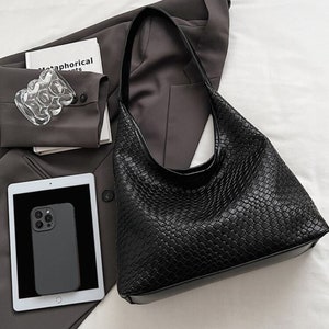 May include: A black woven leather tote bag with a single strap. The bag is on a white surface with a brown blazer and a tablet.