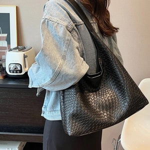 May include: A black woven leather tote bag with a long strap. The bag is worn over the shoulder of a person wearing a light blue denim jacket and a black skirt.
