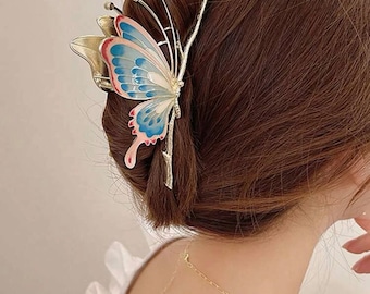 May include: A butterfly-shaped hair clip in gold, blue, and pink. The butterfly design is attached to a gold-coloured metal clip, designed to secure hair. This clip is a fashionable accessory suitable for different hairstyles.