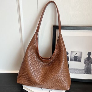 May include: A brown woven leather tote bag with a single top handle. The bag is sitting on a white book with the word "WC" printed on it.
