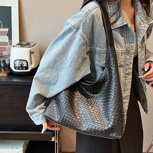 May include: A black woven leather shoulder bag with a long strap. The bag is being worn by a person wearing a light blue denim jacket.