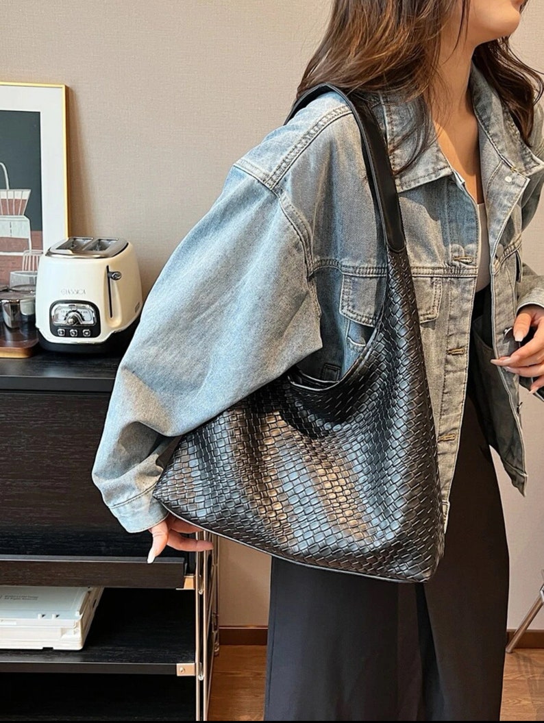 May include: A black woven leather shoulder bag with a long strap. The bag is being worn by a person wearing a light blue denim jacket.