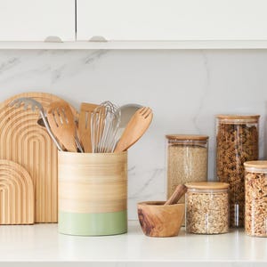 May include: A kitchen countertop with a variety of kitchen utensils and storage containers. A bamboo utensil holder with a green bottom holds wooden spoons, a whisk, and a spatula. There are four glass storage containers with wooden lids filled with various grains and pasta. A wooden mortar and pestle sits on the counter.