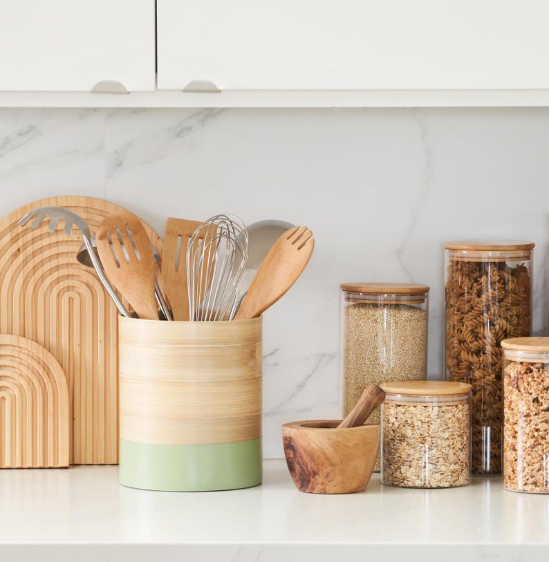 May include: A kitchen countertop with a variety of kitchen utensils and storage containers. A bamboo utensil holder with a green bottom holds wooden spoons, a whisk, and a spatula. There are four glass storage containers with wooden lids filled with various grains and pasta. A wooden mortar and pestle sits on the counter.