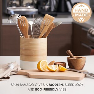 May include: A white and natural bamboo utensil holder with a white base. The holder is filled with various kitchen utensils, including a whisk, wooden spoons, and a spatula. The holder is sitting on a wooden cutting board with lemons and a wooden mortar and pestle.