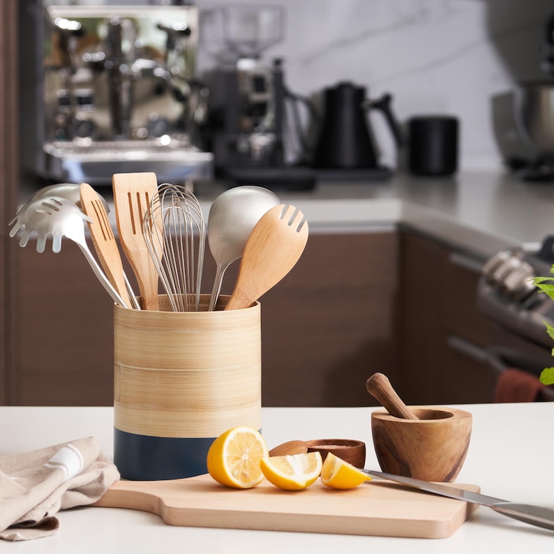 May include: A bamboo utensil holder with a blue stripe, filled with various kitchen utensils, including a whisk, wooden spoons, and a metal ladle. The holder is on a white countertop next to a wooden cutting board with lemon slices.
