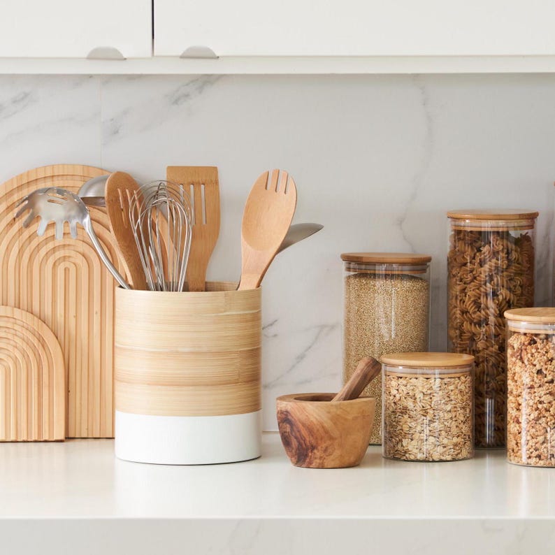 May include: A white and natural bamboo kitchen utensil holder with a variety of wooden spoons, spatulas, and whisks inside.  There are also several glass jars with lids on a marble countertop, filled with various grains and pasta.