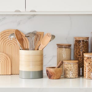 May include: A bamboo utensil holder with a gray base, filled with wooden spoons, a fork, a ladle, and a whisk. The holder is on a white countertop next to a wooden mortar and pestle and four glass jars with lids, filled with various grains and cereals.